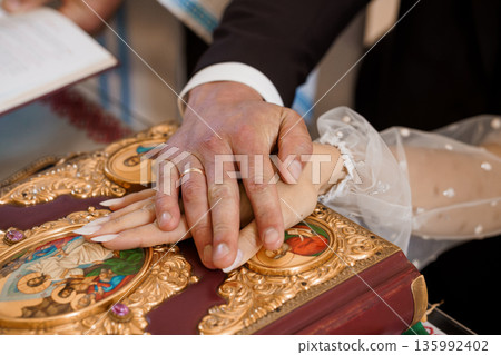 Orthodox Wedding Ceremony Hands Holding Holy Book with Golden Icons and Ring Exchange in Church Setting 135992402