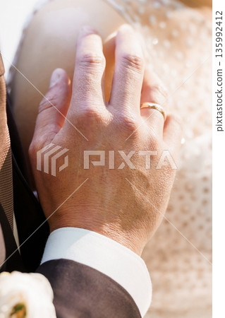 Close-up of Groom's Hand Holding Bride's Shoulder in Wedding Ceremony with Gold Ring and Lace Dress 135992412