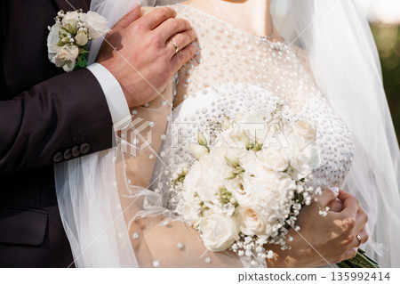 Close-up of a wedding couple embracing, bride in white beaded gown holding bouquet, groom in dark suit with boutonniere, soft natural lighting 135992414