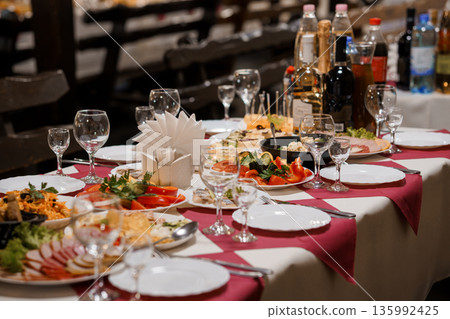 Elegant Banquet Table Setup with Assorted Delicacies, Wine Bottles, and Fine Glassware on Red and White Checkered Tablecloth Elegant Banquet Table Setup with Assorted Delicacies, Wine Bottles, and Fine Glassware on Red and White Checkered Tablecloth 135992425