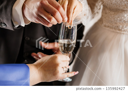 Close-up of bride and groom placing wedding rings into champagne glass during intimate ceremony moment 135992427