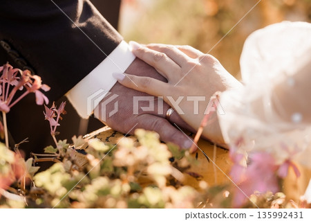 Close-up of a bride and groom holding hands with wedding rings, soft sunlight, pink flowers, and elegant white lace fabric in a romantic outdoor setting 135992431