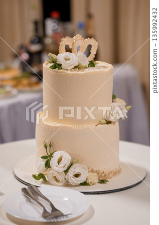 Elegant Two-Tier Wedding Cake with White Roses and Gold Heart Decorations on Cream Fondant Background 135992432