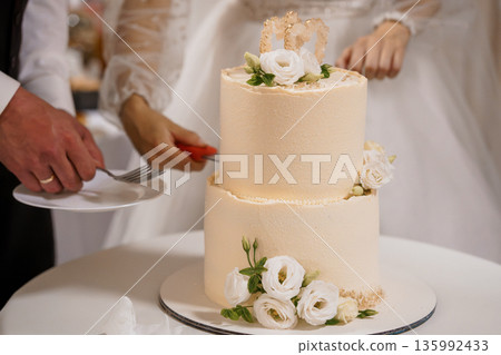 Elegant Two-Tier Wedding Cake with White Roses and Gold Heart Decorations Being Sliced by Bride and Groom 135992433