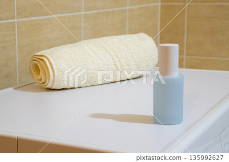 Rolled beige towel and blue bottle with plastic cap on washing machine top. Laundry or bathroom concept. Cleanliness and hygiene idea. Rolled beige towel and blue bottle with plastic cap on washing machine top. Laundry or bathroom concept. Cleanliness and hygiene idea. 135992627