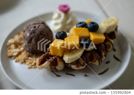 Waffle with chocolate ice cream served with sliced banana and mango, whipped cream and sauce. 135992804