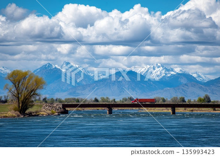 Red Truck Driving Across a Bridge with Dramatic Clouds and Snowy Mountains in Background 135993423