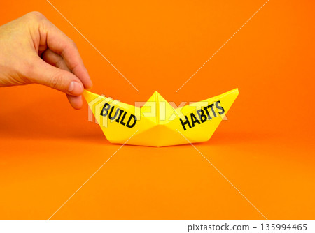 Build habits symbol. Concept word Build habits on beautiful yellow paper boat. Businessman's hand. Beautiful orange paper background. Business build habits concept. Copy space. Build habits symbol. Concept word Build habits on beautiful yellow paper boat. Businessman's hand. Beautiful orange paper background. Business build habits concept. Copy space. 135994465