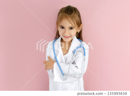 Smiling little girl wearing doctor coat and stethoscope, dreaming of medical career. Concept of healthcare education, International Day, childhood ambition and future profession. Smiling little girl wearing doctor coat and stethoscope, dreaming of medical career. Concept of healthcare education, International Day, childhood ambition and future profession. 135995639