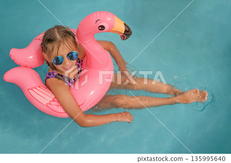 Smiling child relaxing on pink flamingo inflatable in clear blue swimming pool. Summer vacation, pool fun, kids leisure, tropical holiday mood and joyful childhood concept. Smiling child relaxing on pink flamingo inflatable in clear blue swimming pool. Summer vacation, pool fun, kids leisure, tropical holiday mood and joyful childhood concept. 135995640