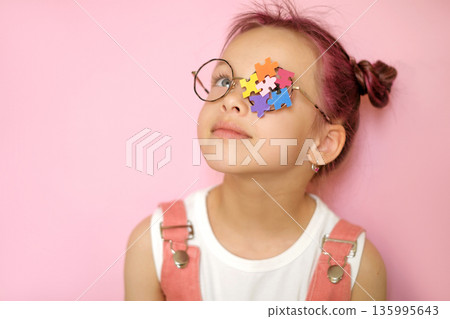 Young girl wearing glasses with colorful puzzle pieces covering one eye. Concept of autism awareness, neurodiversity, child development, education and inclusion on pink background. 135995643