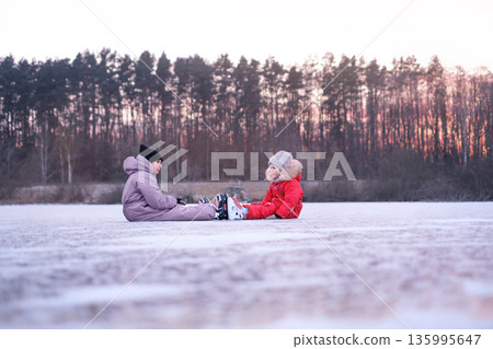 Two children resting on frozen lake after ice skating. Winter outdoor activity, childhood leisure, safety moment, playful pause during cold seasonal recreation in nature. Two children resting on frozen lake after ice skating. Winter outdoor activity, childhood leisure, safety moment, playful pause during cold seasonal recreation in nature. 135995647