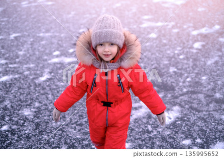 Cute child in bright red winter snowsuit standing on frozen ice outdoors. Winter season, cold weather, childhood lifestyle, outdoor activity and safe winter recreation. Cute child in bright red winter snowsuit standing on frozen ice outdoors. Winter season, cold weather, childhood lifestyle, outdoor activity and safe winter recreation. 135995652