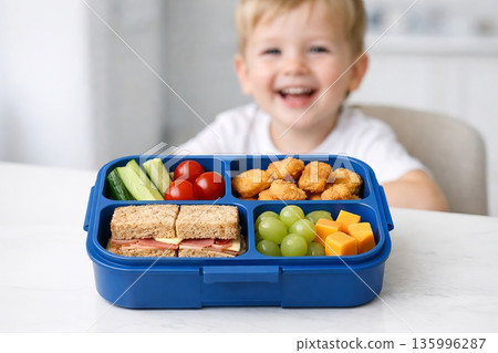 Happy blond boy with blue lunchbox containing healthy and delicious food. Kids school meal. Healthy eating concept for children 135996287