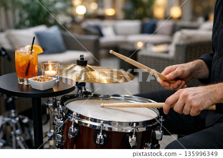 Man playing drums with drumsticks in a cozy indoor setting. Evening entertainment and live music concept for hospitality. Vibrant cocktail, candle 135996349