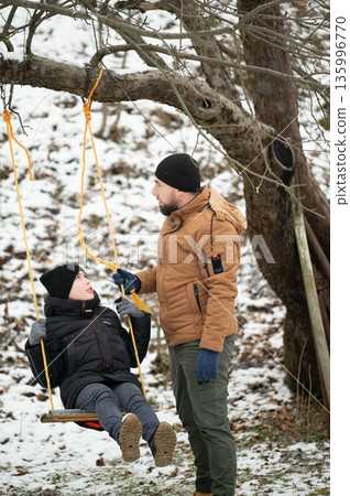 A father gently pushes his son on a rope swing in a snowy winter landscape. A heartwarming moment of shared outdoor play. A father gently pushes his son on a rope swing in a snowy winter landscape. A heartwarming moment of shared outdoor play. 135996770