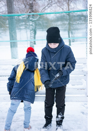 Two children play in the snow, one carefully crafting a snowball while the other watches. Two children play in the snow, one carefully crafting a snowball while the other watches. 135996824