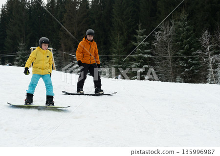 Two enthusiastic young snowboarders are having a fantastic winter adventure as they glide down a snowy slope, surrounded by towering, majestic evergreen trees that create a breathtaking backdrop Two enthusiastic young snowboarders are having a fantastic winter adventure as they glide down a snowy slope, surrounded by towering, majestic evergreen trees that create a breathtaking backdrop 135996987