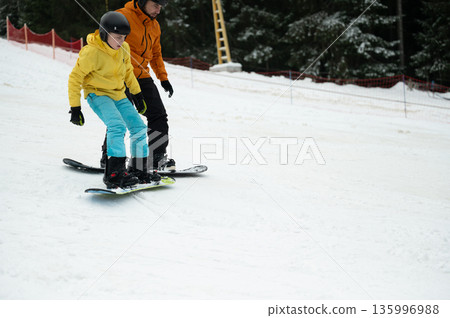 Snowboarders gliding down a snowy slope on a winter afternoon near a ski resort surrounded by tall pine trees and fresh powder Snowboarders gliding down a snowy slope on a winter afternoon near a ski resort surrounded by tall pine trees and fresh powder 135996988