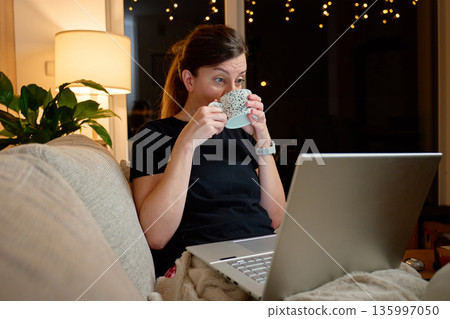 Adult woman sitting on sofa at home drinking from mug while using laptop at night 135997050