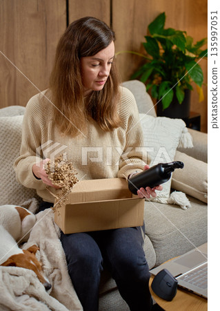 Woman sitting on sofa opening cardboard box and examining reusable bottle. Concept of online shopping 135997051