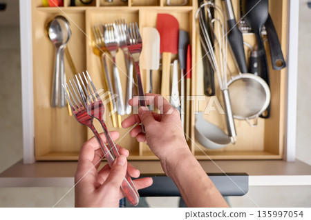 Hands holding forks above kitchen drawer with cooking utensils. Concept of kitchen organization Hands holding forks above kitchen drawer with cooking utensils. Concept of kitchen organization 135997054