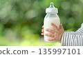 Mother or caregiver feeding infant with bottle in natural daylight at outdoor setting 135997101