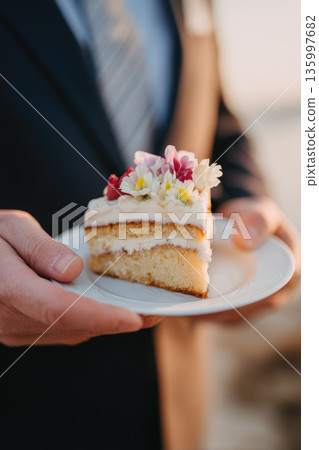 Vegan wedding cake slice with creamy frosting and fresh flowers held by person in formal attire at outdoor celebration, creating joyful and elegant atmosphere 135997682
