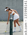 Female tennis player in white sportswear picking up ball on modern outdoor court during training 135998141