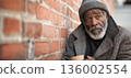 An elderly African American man with a gray beard sits against a worn brick wall. He appears lost in thought, holding a small cup beside him filled with coins 136002554