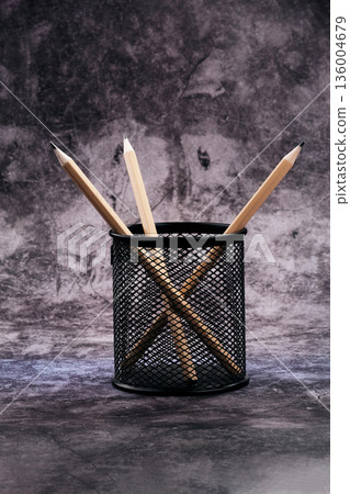 Three light wood sharpened pencils stand neatly in a black metal mesh cup on a mottled dark gray background 136004679