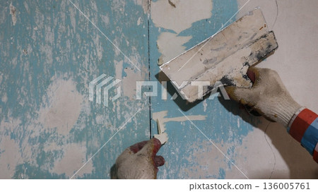 Hands in dirty gloves holding an old wide spatula and a piece of torn wallpaper against a peeling wall, scraping off old wallpaper and glue residue with a trowel, preparing the wall for renovation 136005761
