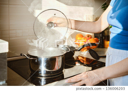 Unrecognizable woman lifts the lid of a pot of boiling water 136006203