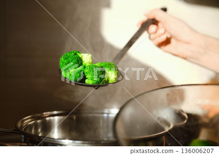A person holds a slotted spoon with steamed broccoli above a pot in a kitchen 136006207