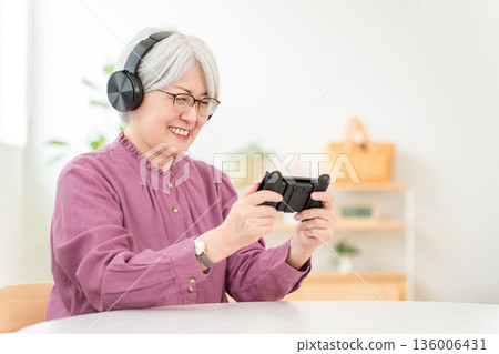 一位頭髮花白的亞裔女性戴著耳機，在家裡的客廳玩遊戲。 136006431