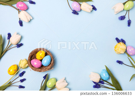 Easter colored painted eggs in bird nest with yellow daffodils, white tulips, and blue muscari on blue background with space for text and greetings. Easter and spring concept. Spring background 136006633