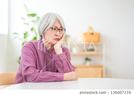 A silver-haired senior woman sitting at the dining table in her home and thinking 136007239