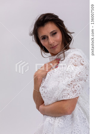 Woman posing in white lace garment with arm across body and subtle smile, refined styling and delicate texture, fashionforward yet intimate mood on studio backdrop 136007509