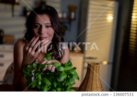 Herb care evening, Serene woman delicately tending aromatic basil under soft lighting, Gentle scene of woman caring for fragrant basil in cozy nighttime kitchen environment 136007531