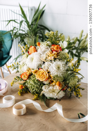 Mixed floral bouquet sits on craft paper next to ribbon and tape on table. Locally grown flowers, seasonal floristry, eco-friendly flower shop. Mixed floral bouquet sits on craft paper next to ribbon and tape on table. Locally grown flowers, seasonal floristry, eco-friendly flower shop. 136007736