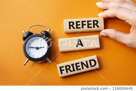 Refer a Friend symbol. Concept words Refer a Friend on wooden blocks. Businessman hand. Beautiful orange background with alarm clock. Business and Refer a Friend concept. Copy space. 136008448