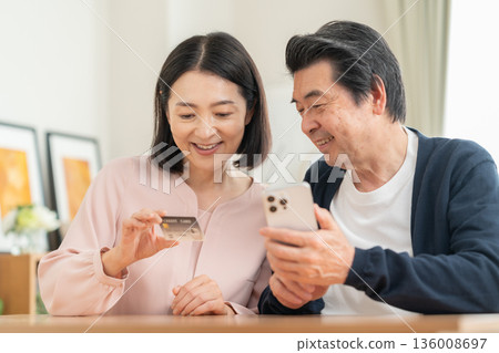 A middle-aged couple looking at their smartphone and credit card in the living room A middle-aged couple looking at their smartphone and credit card in the living room 136008697