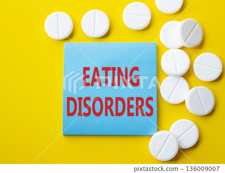 Eating Disorders symbol. Concept word Eating Disorders on blue note. Beautiful yellow background with pills. Medicine and Eating Disorders concept. Copy space Eating Disorders symbol. Concept word Eating Disorders on blue note. Beautiful yellow background with pills. Medicine and Eating Disorders concept. Copy space 136009007