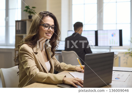 Happy businesswoman using laptop and pencil at bright office workstation and graphs Happy businesswoman using laptop and pencil at bright office workstation and graphs 136009046