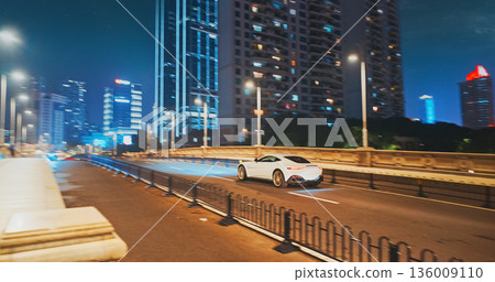 White supercar in fast motion at night. urban street scene with moving cars and modern architecture illuminated at night 136009110