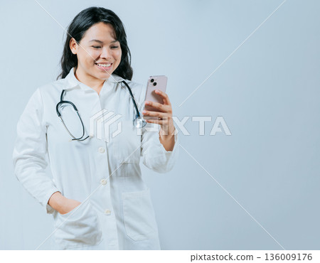 Female healthcare worker holding cellphone. Smiling asian doctor using smartphone isolated, Young asian female doctor using phone 136009176