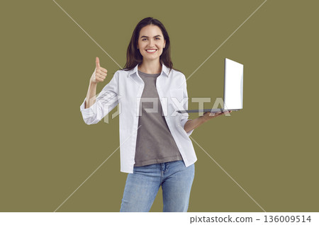 Smiling brunette woman in casual clothes showing thumb up holding laptop on khaki background. Smiling brunette woman in casual clothes showing thumb up holding laptop on khaki background. 136009514