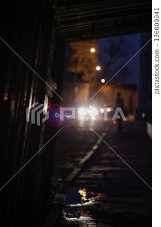 Silhouette in rainy night alley with car lights bokeh, moody urban scene. Man walking in dark passage with glowing headlights and reflections. Night City Street View Silhouette in rainy night alley with car lights bokeh, moody urban scene. Man walking in dark passage with glowing headlights and reflections. Night City Street View 136009941