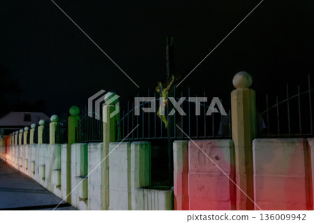 Crucifix Behind Fence at Night. A wooden crucifix with a figure of Jesus Christ standing behind a concrete and metal picket fence. Easter faith symbol on vibrant fence under starry sky Crucifix Behind Fence at Night. A wooden crucifix with a figure of Jesus Christ standing behind a concrete and metal picket fence. Easter faith symbol on vibrant fence under starry sky 136009942