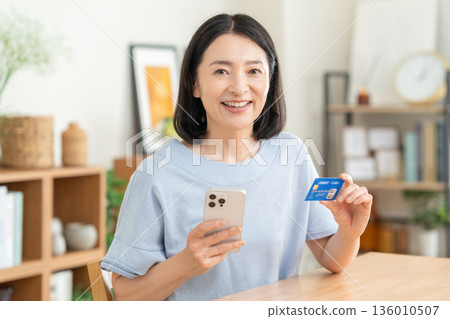 A middle-aged woman looking at her credit card and smartphone in the living room A middle-aged woman looking at her credit card and smartphone in the living room 136010507